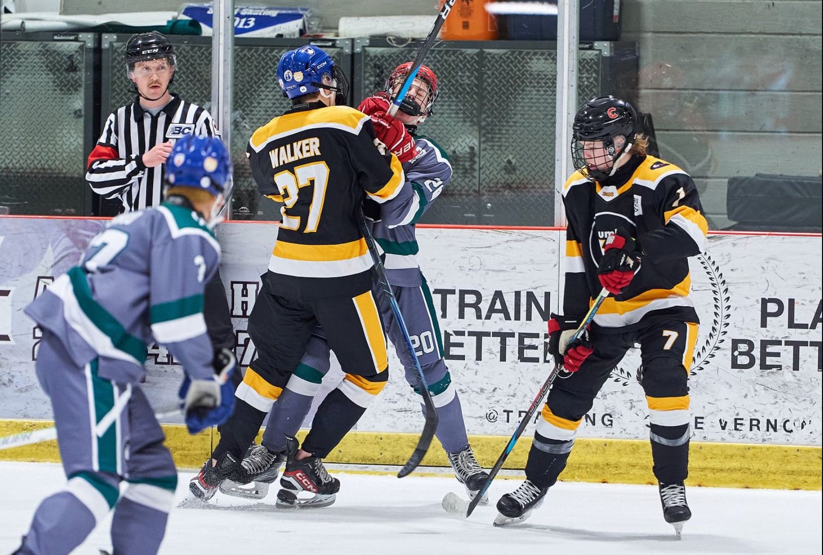 Black-and-gold and grey-and-teal players beside the boards with a referee at UMH-68.