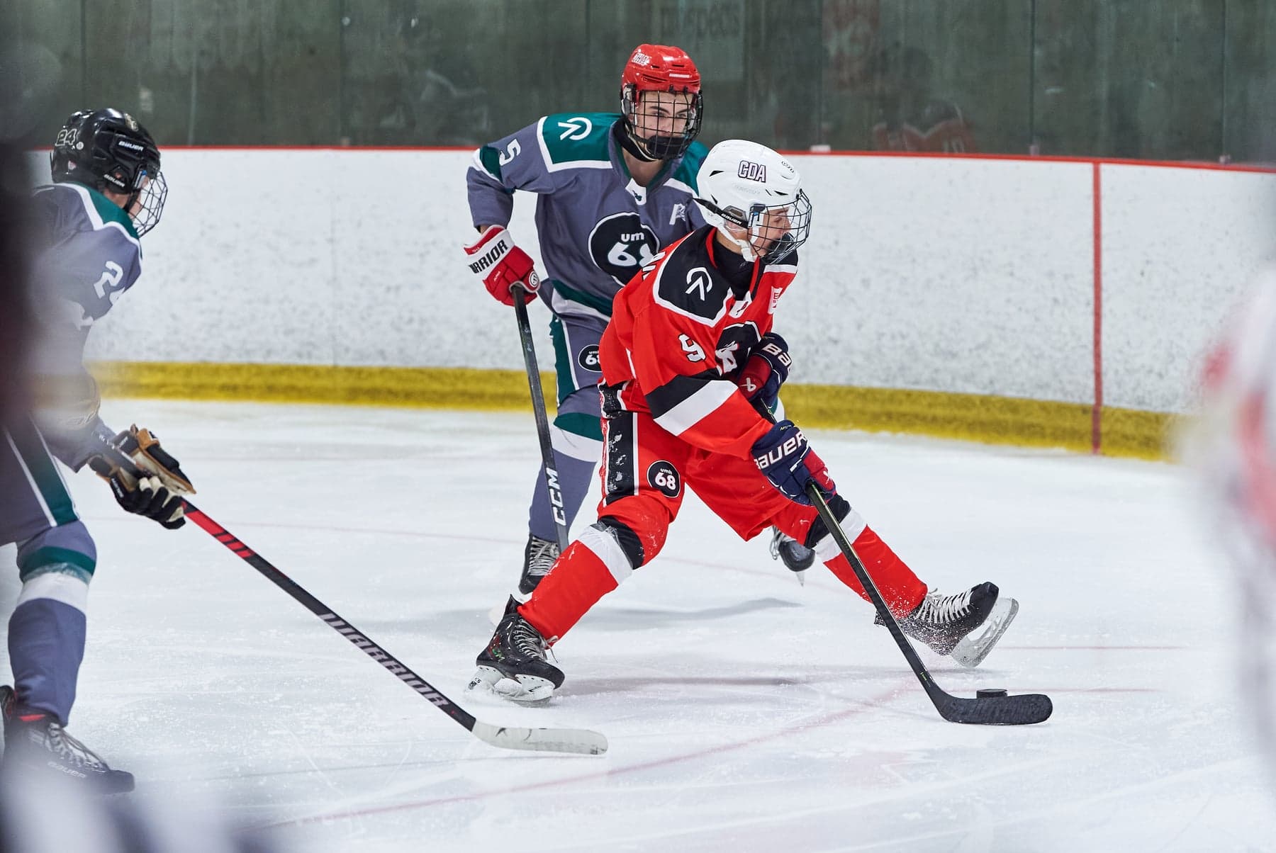 A red-jersey player skates between grey-and-teal opponents during a UMH-68 game.