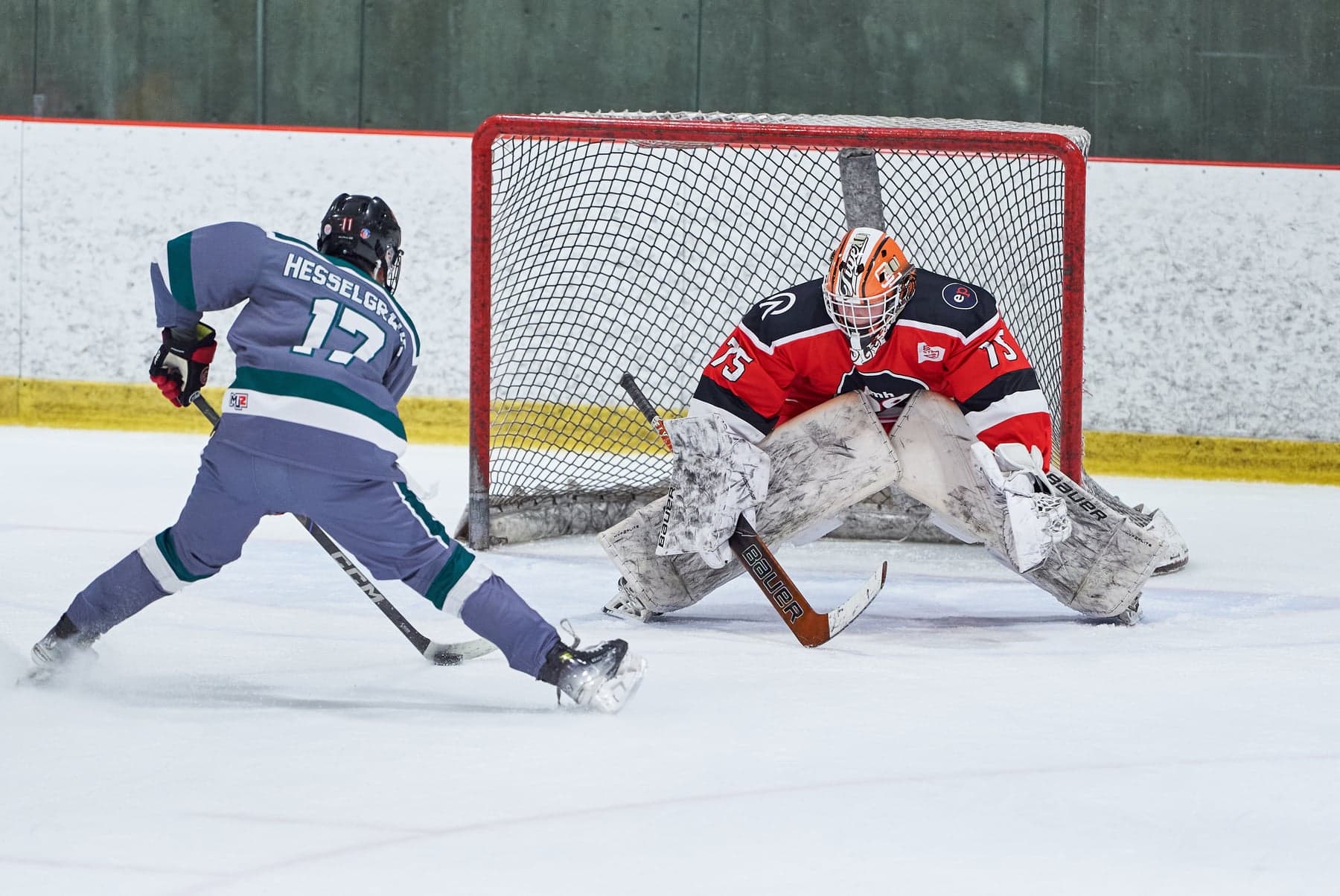 A red-jersey goalie faces a shot from a grey-and-teal skater near the net.