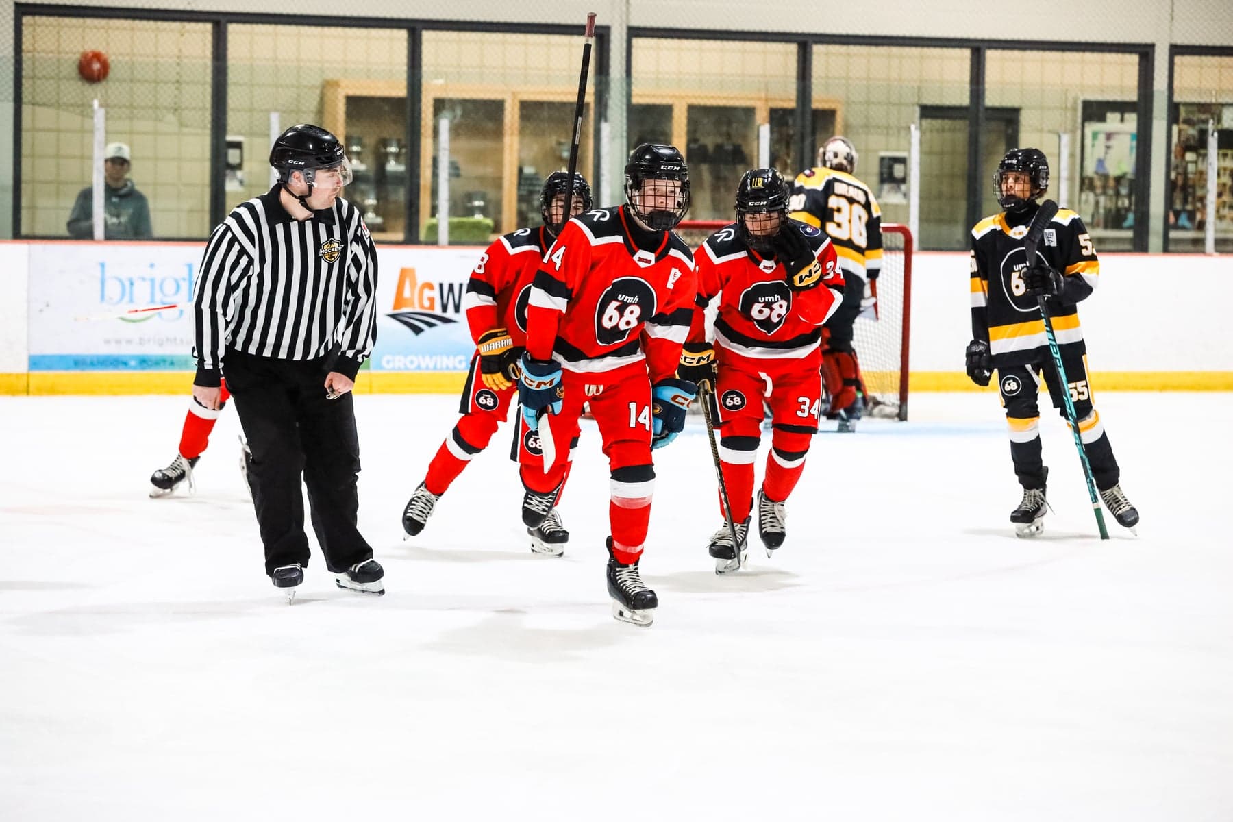 Red-jersey UMH-68 teammates skate beside a referee with black-and-gold opponents near the net.