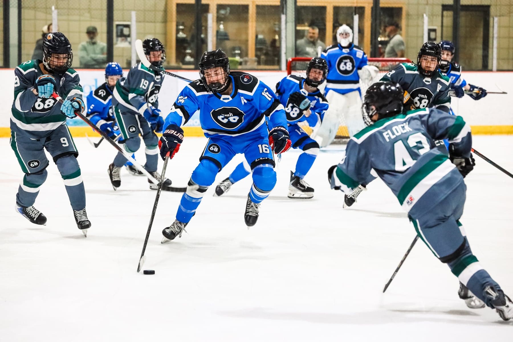 A blue-jersey player carries the puck through grey-and-teal opponents during a UMH-68 game.