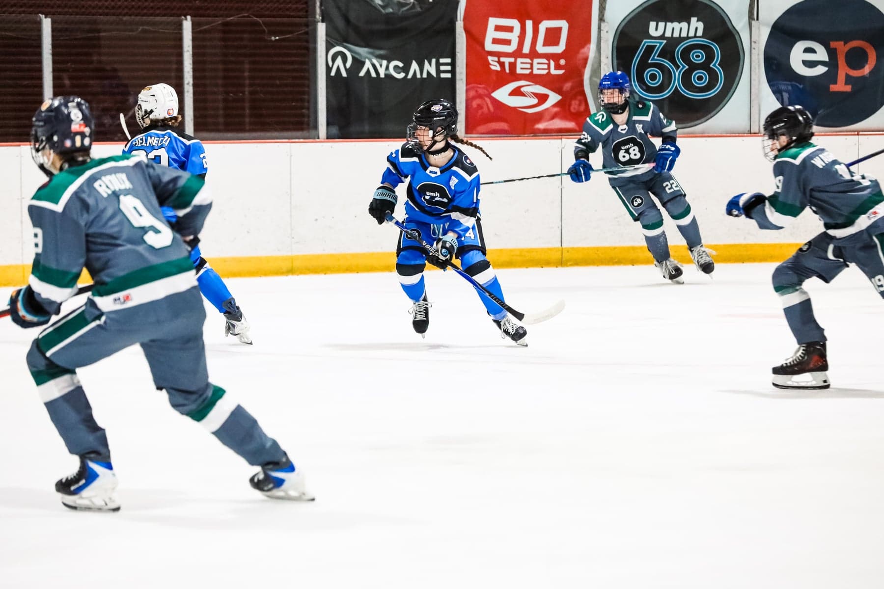Blue and grey-and-teal players follow the play in front of UMH-68 and sponsor banners.