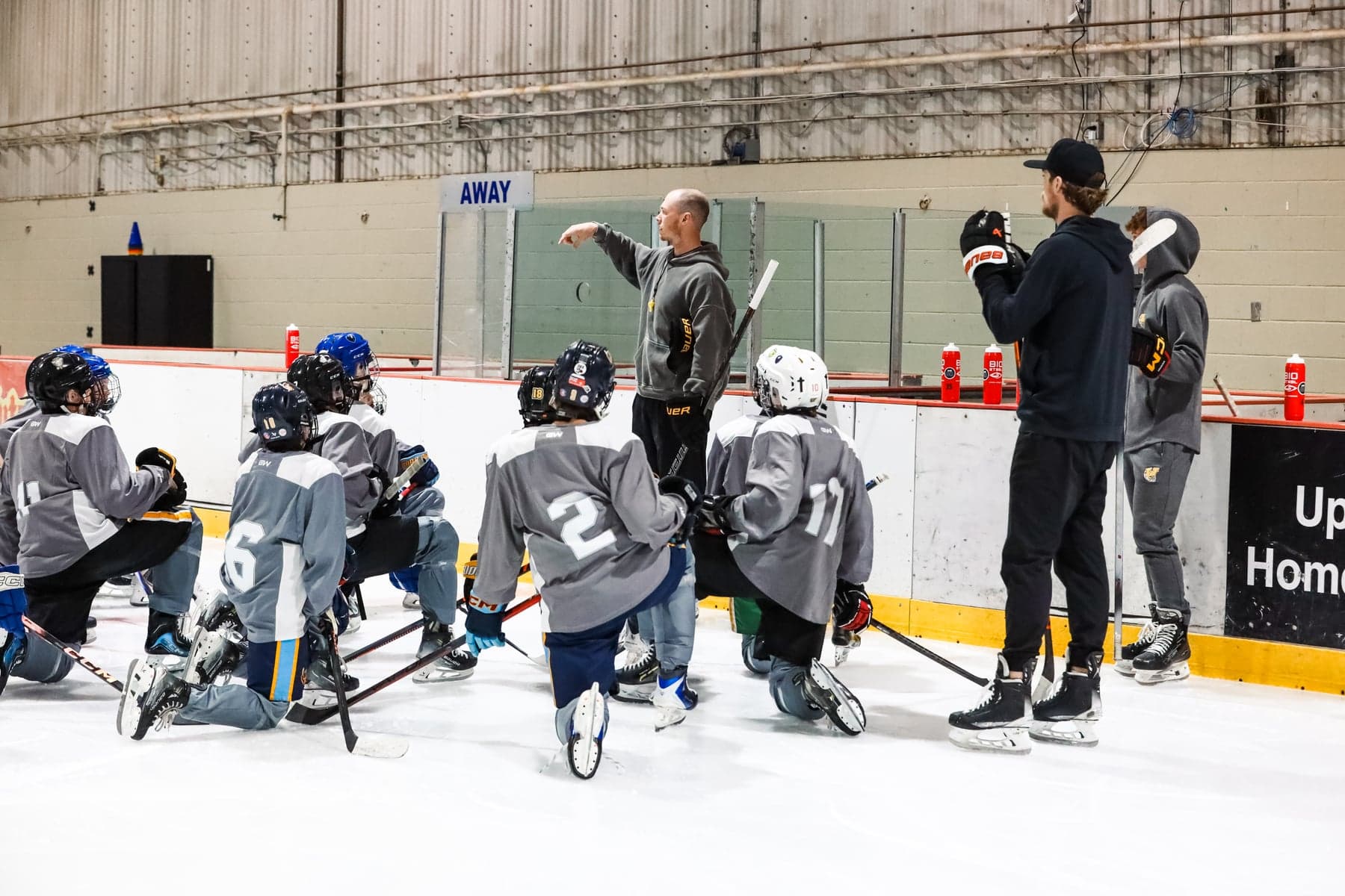 Coaches lead an on-ice session as grey-jersey players kneel and listen beside the boards.