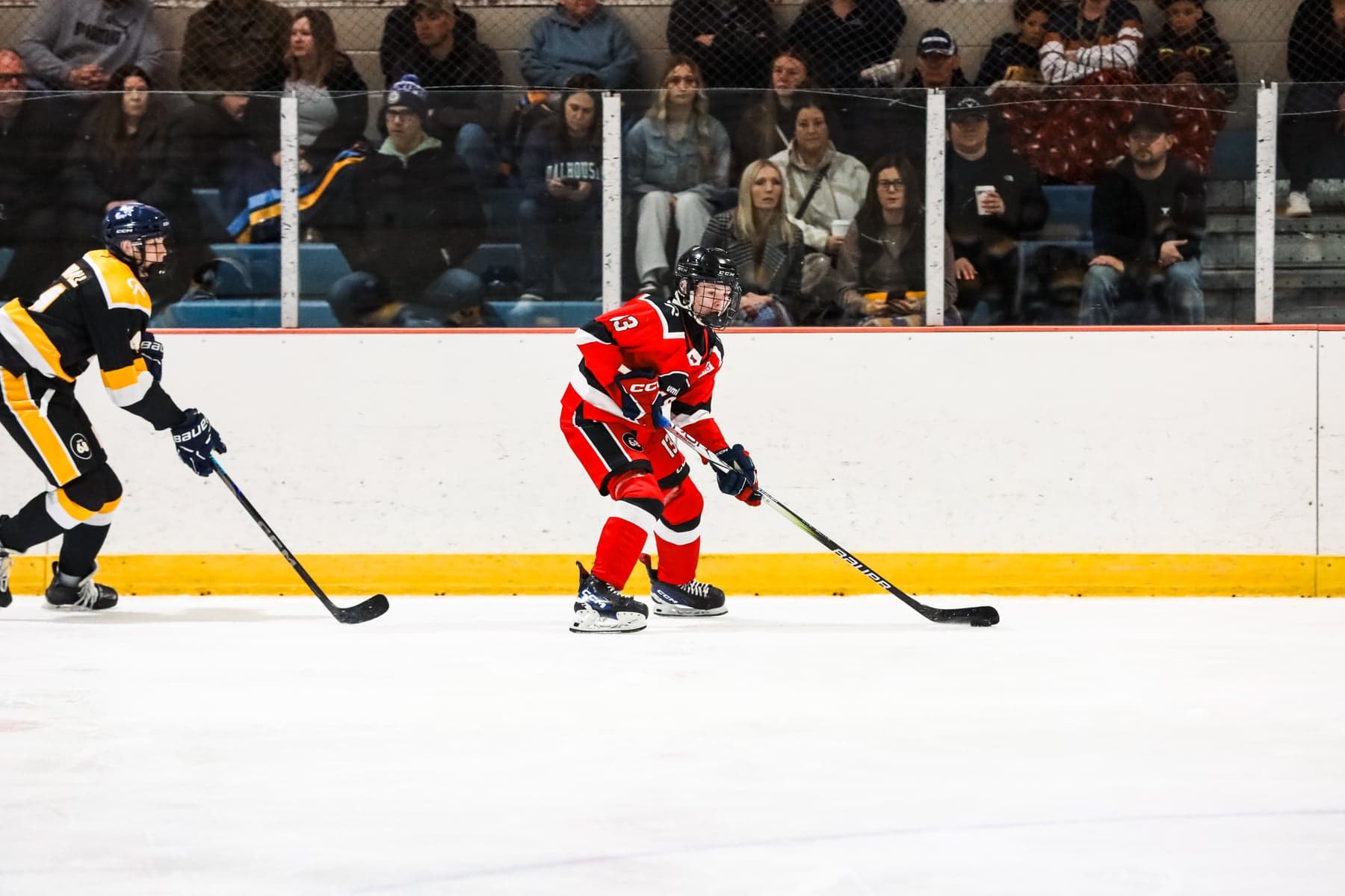 A red-jersey player skates with the puck along the boards as spectators watch from the stands.