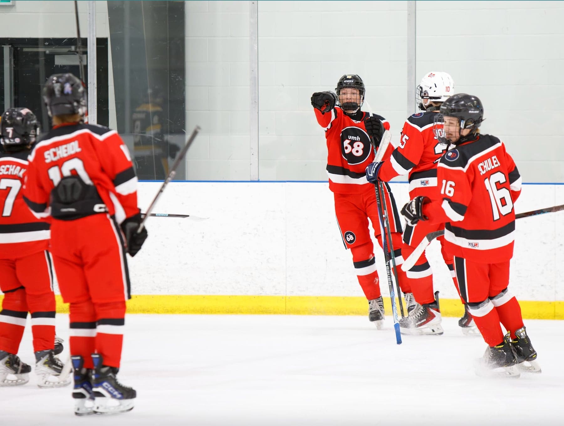 Red-jersey UMH-68 teammates gather beside the boards as a player points toward the rink.