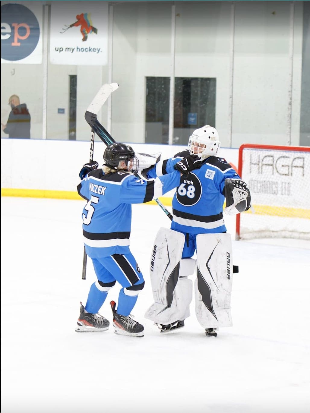 A blue-jersey skater and goalie celebrate together in front of the net.