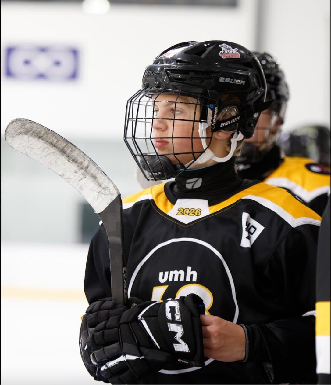 A black-and-gold UMH-68 player watches the rink while holding his hockey stick.
